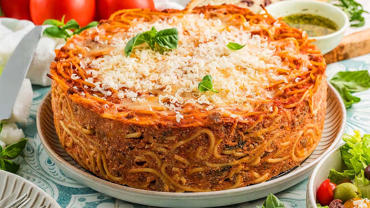 A round baked spaghetti pie topped with grated cheese and basil leaves, served on a plate with fresh vegetables and sauce in the background.