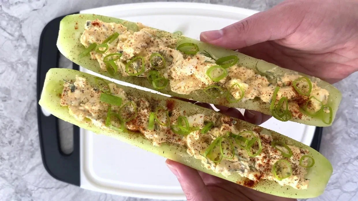 Two cucumber halves filled with an egg salad mixture, topped with sliced green onions and sprinkled with seasoning, held over a white cutting board.