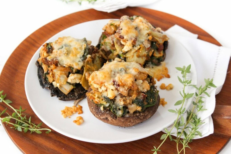 Three stuffed portobello mushrooms filled with a vegetable and cheese mixture are served on a white plate with a wooden base, garnished with fresh herbs.