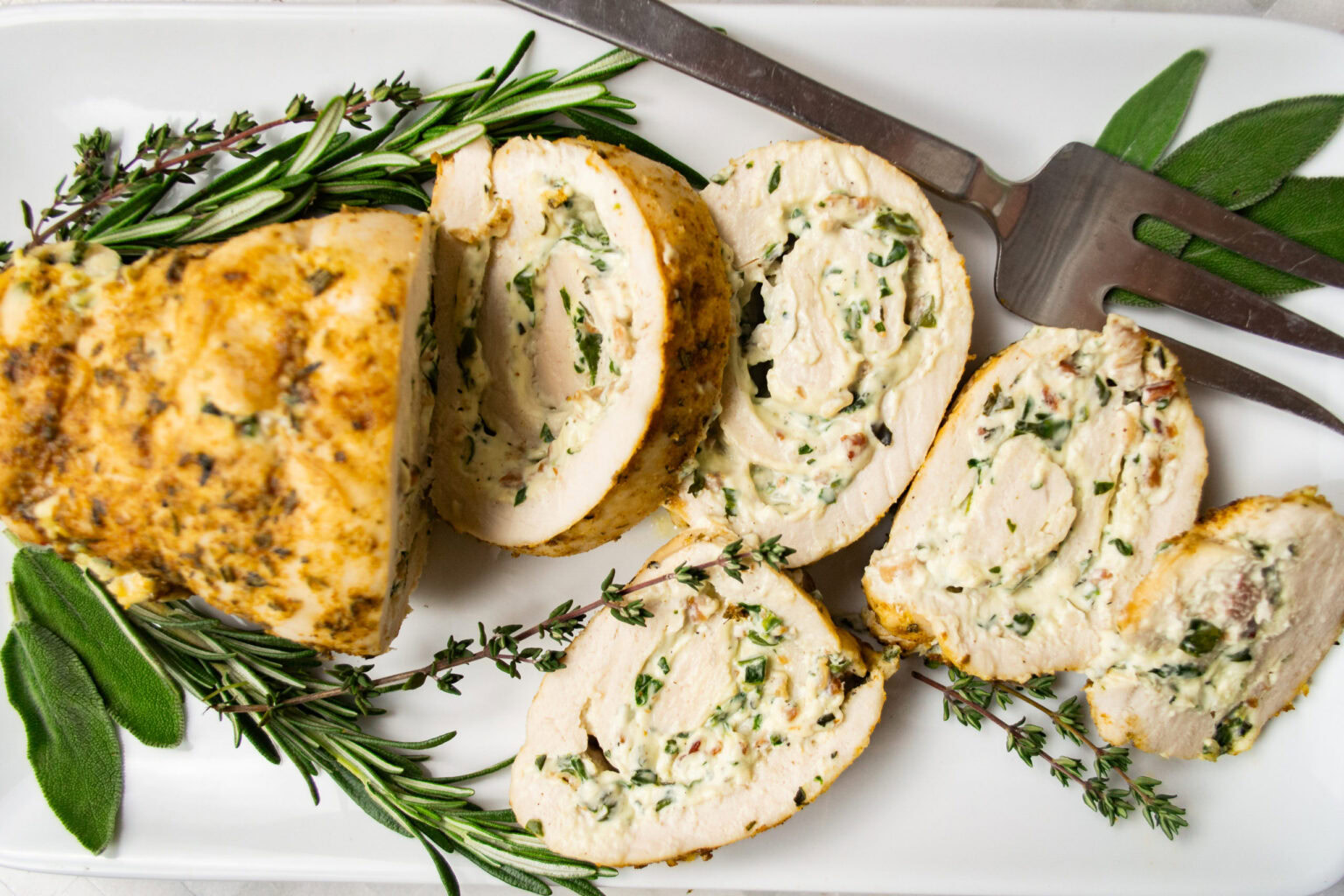 Sliced stuffed chicken breast filled with herbs and cheese, garnished with sprigs of rosemary and thyme on a white platter with serving utensils.