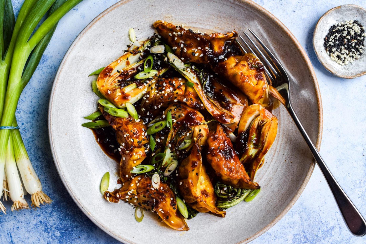 A plate of pan-fried dumplings topped with sliced green onions and sesame seeds, with a fork on the side; green onions and a small dish of sesame seeds are nearby.