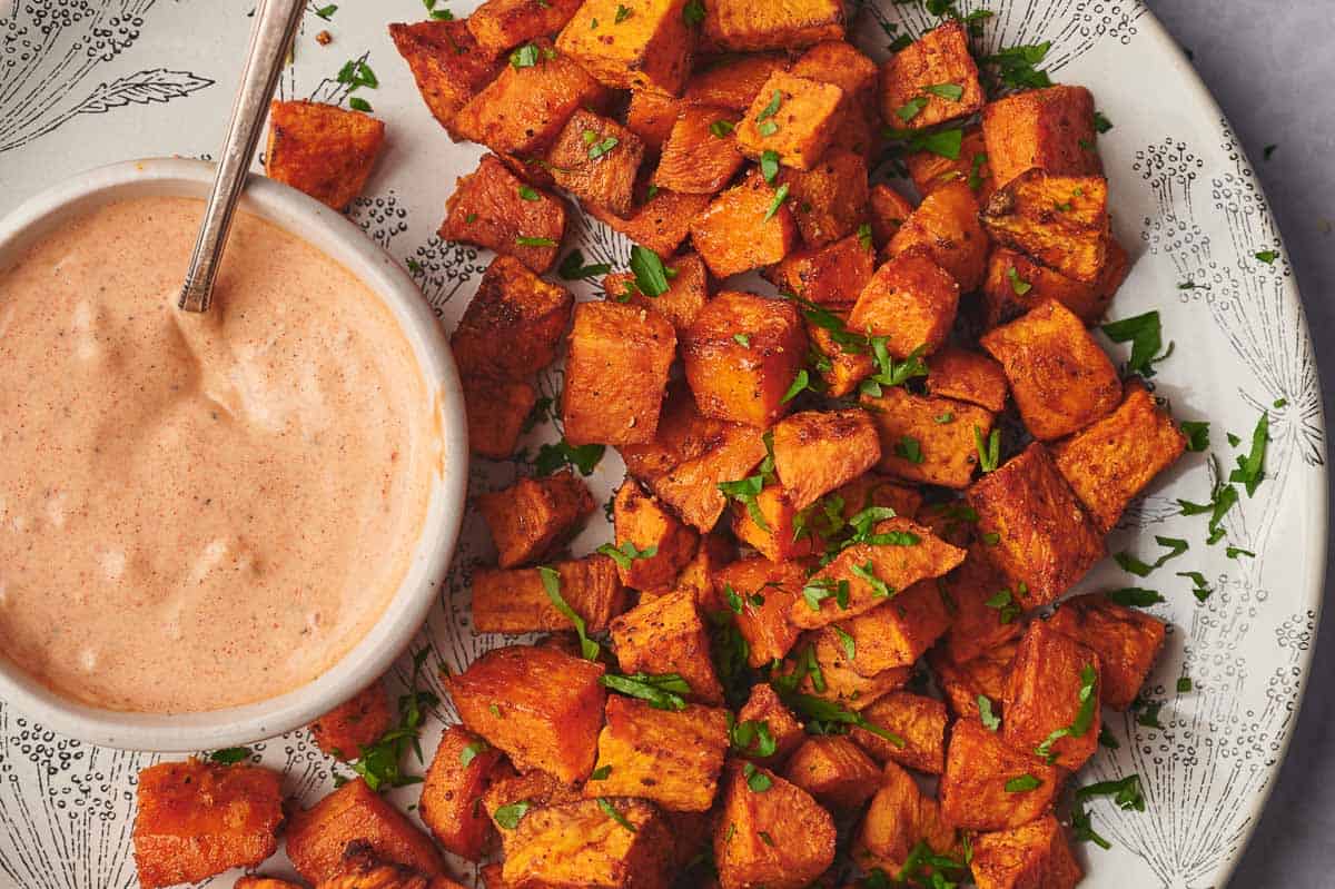 A plate of roasted sweet potato cubes garnished with chopped herbs, served with a bowl of creamy dipping sauce.