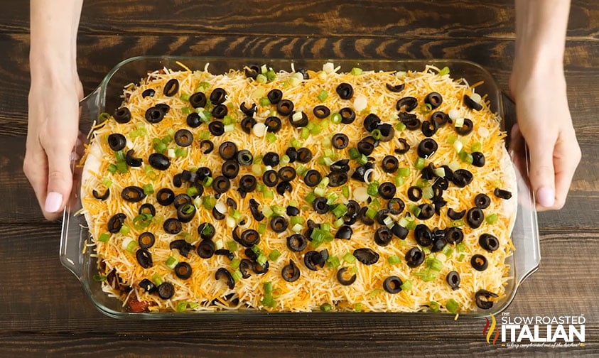 A person holds a glass dish filled with layered dip topped with shredded cheese, sliced black olives, and chopped green onions on a wooden table.
