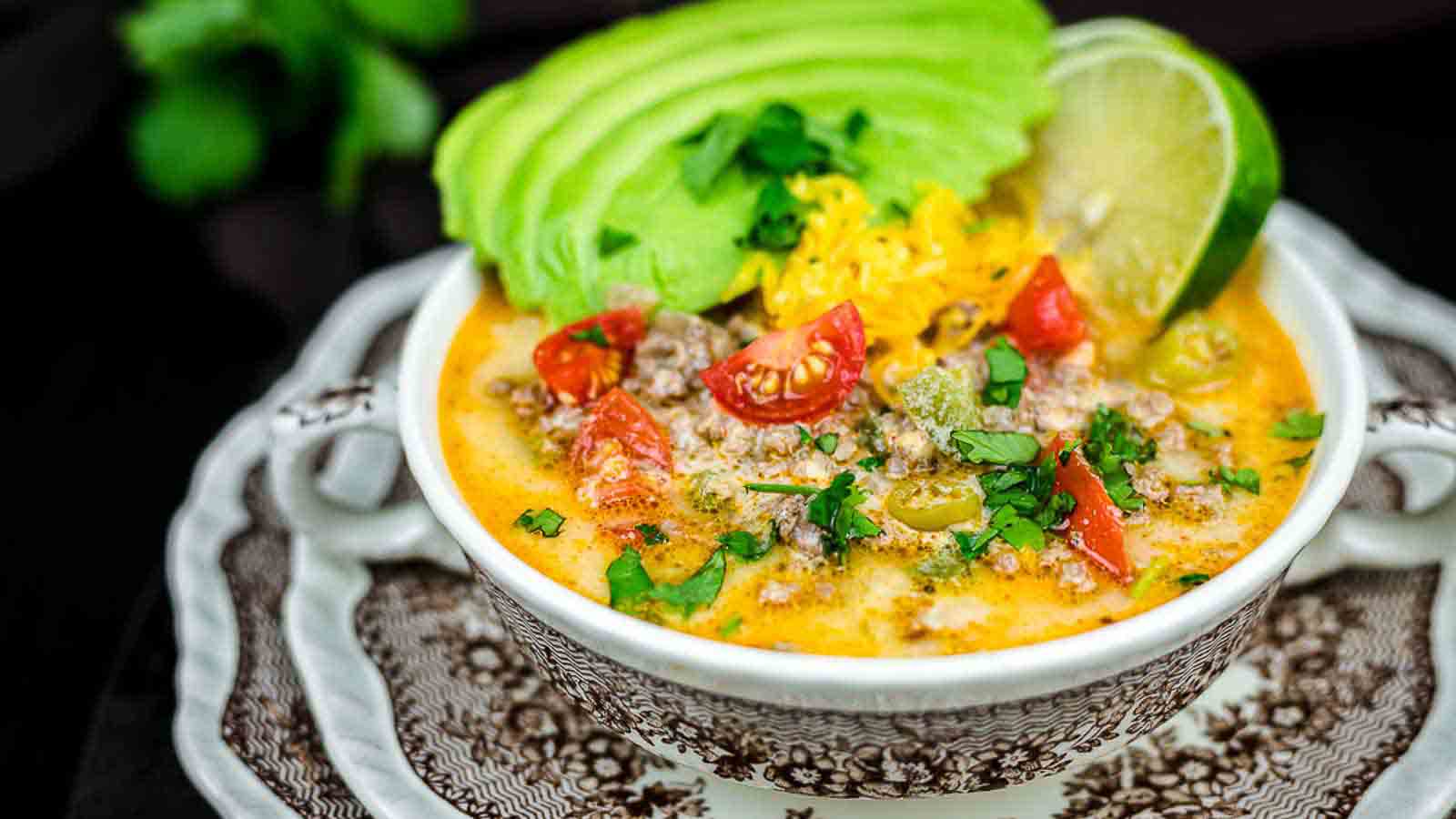 A bowl of creamy soup topped with sliced avocado, shredded cheese, chopped tomatoes, cilantro, and a lime wedge, served on a decorative saucer.