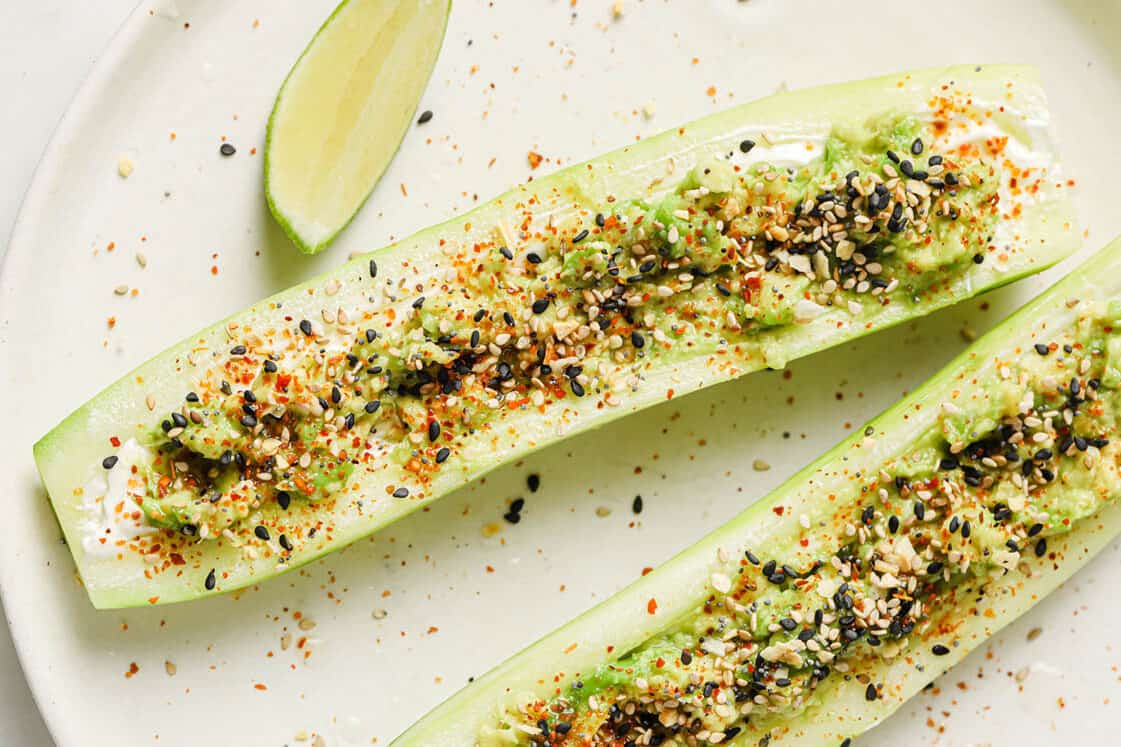 Two cucumber halves topped with a creamy spread, seasoning, and seeds, served on a white plate with a lime wedge.