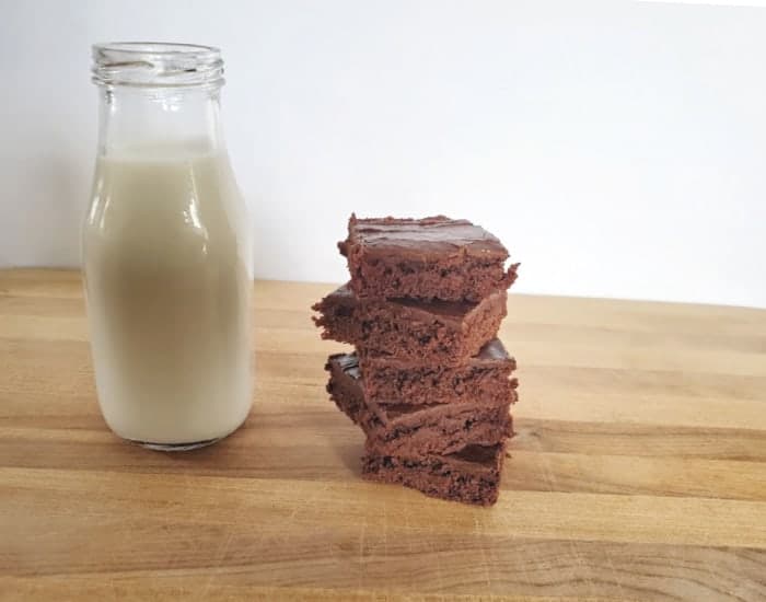 A glass bottle of milk stands next to a stack of five chocolate brownies on a wooden surface with a plain background.