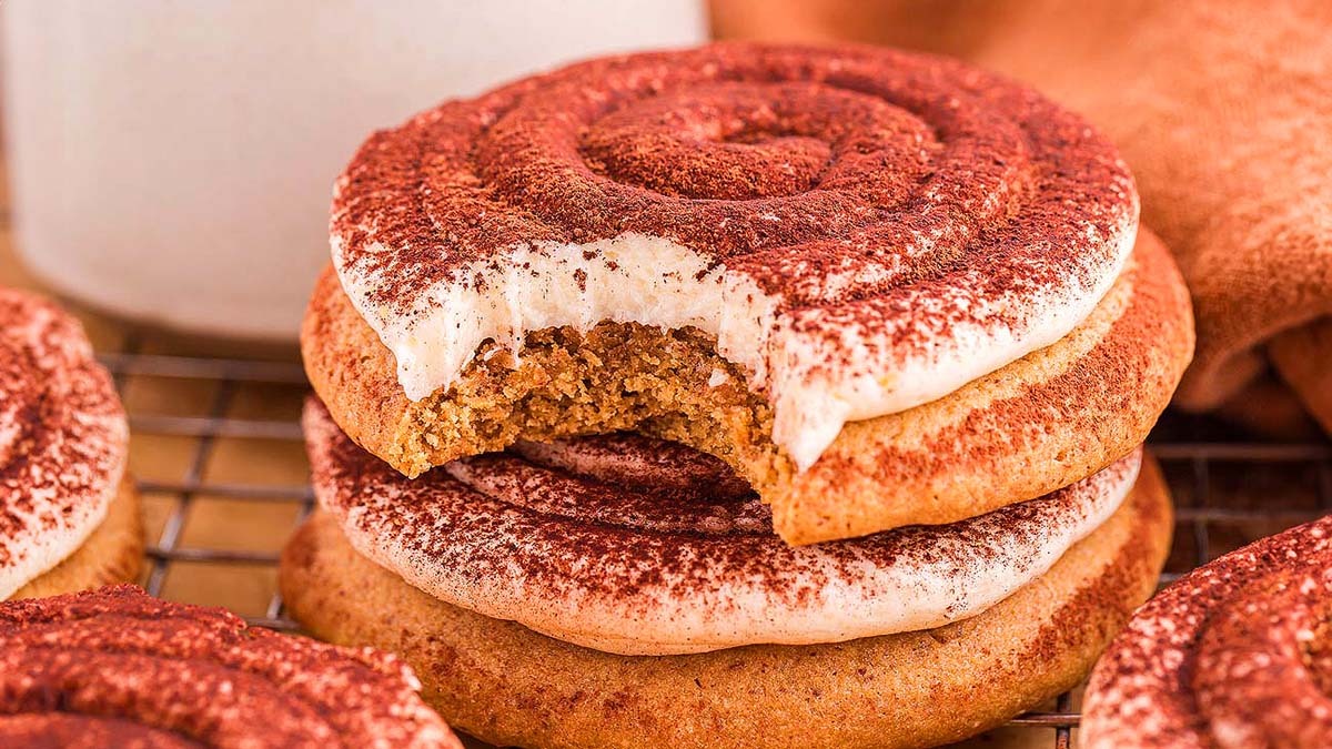 A stack of three frosted cookies dusted with cocoa powder, with a bite taken out of the top cookie, placed on a cooling rack.