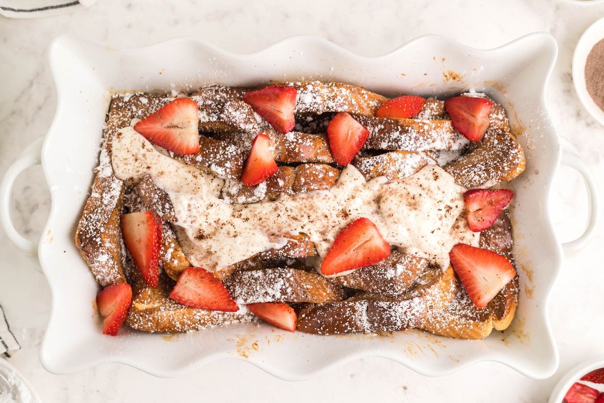 A white baking dish with baked French toast topped with powdered sugar, whipped cream, cinnamon, and sliced strawberries on a marble surface.