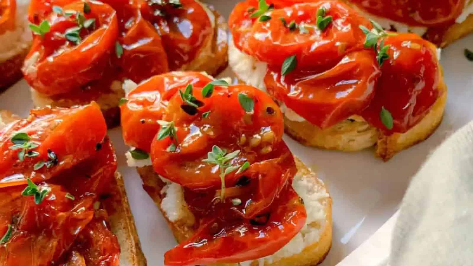 Slices of toasted bread topped with creamy cheese, roasted cherry tomatoes, and fresh green herbs are arranged on a white plate.