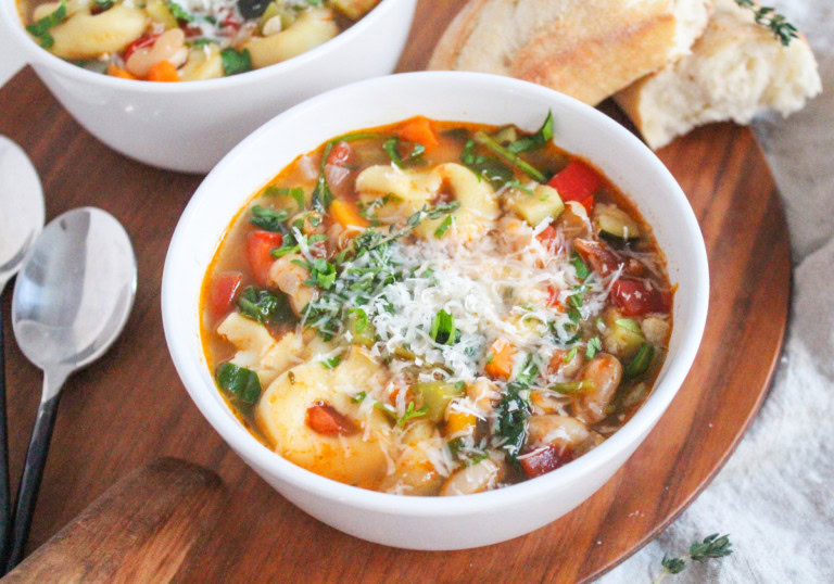 Two bowls of vegetable soup with pasta, topped with grated cheese and herbs, are served on a wooden board with two spoons and pieces of crusty bread on the side.