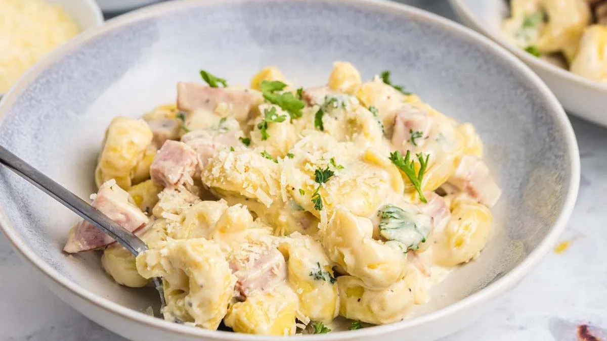A bowl of creamy pasta with ham, cheese sauce, and chopped parsley, served with a spoon on a light-colored table.