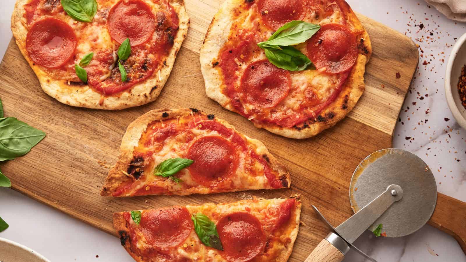 Four small pepperoni pizzas with basil leaves on a wooden cutting board, one sliced, next to a pizza cutter and a bowl of red pepper flakes.