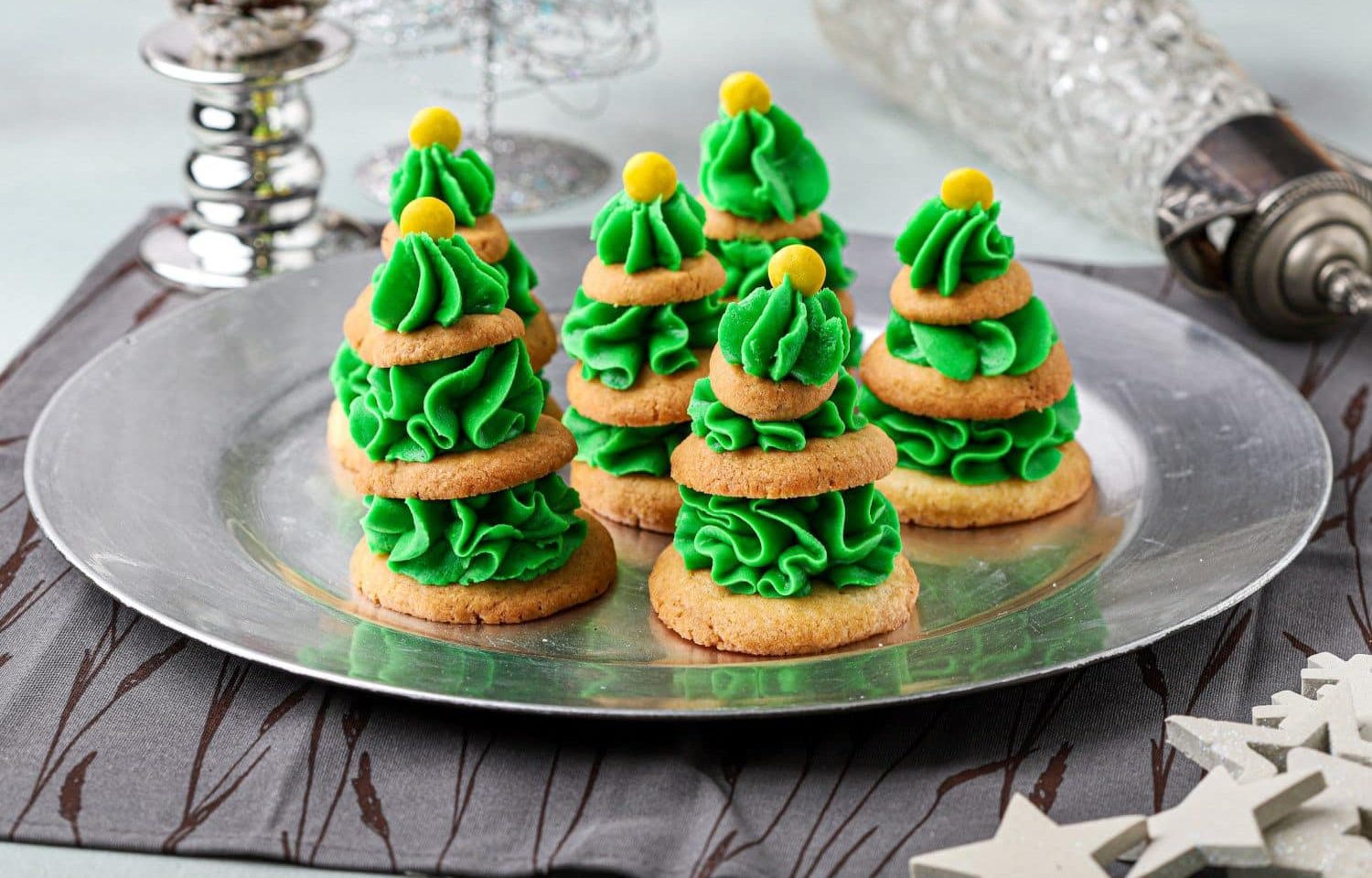 Cookies stacked with green frosting to resemble Christmas trees, each topped with a yellow candy, arranged on a silver plate.