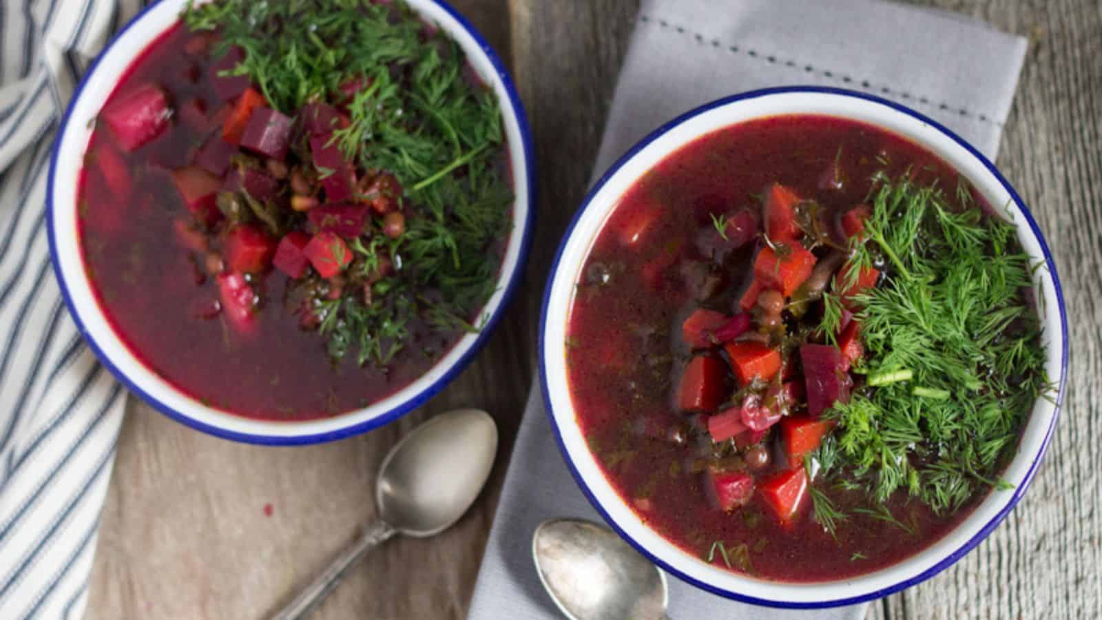 Two bowls of beet soup garnished with fresh dill, placed on a wooden surface beside a striped cloth and two metal spoons.