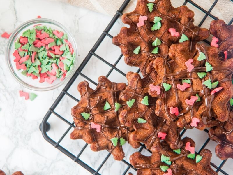 Chocolate waffles with green and red holiday sprinkles are on a cooling rack next to a small glass bowl filled with similar sprinkles.