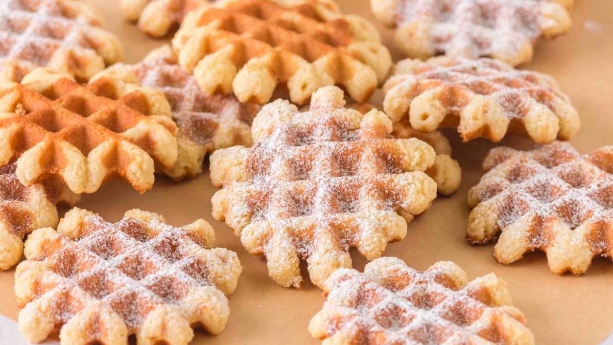 A group of small waffle cookies dusted with powdered sugar, arranged on a light brown surface.