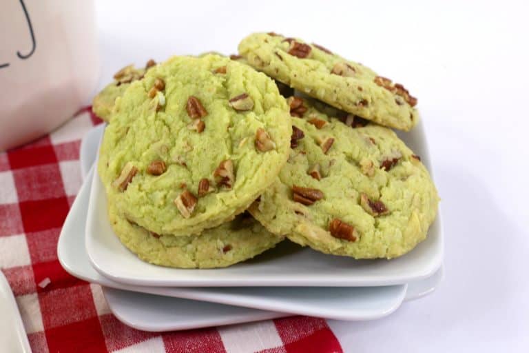 A plate of green cookies topped with chopped pecans is displayed on stacked white dishes, set on a red and white checkered cloth.