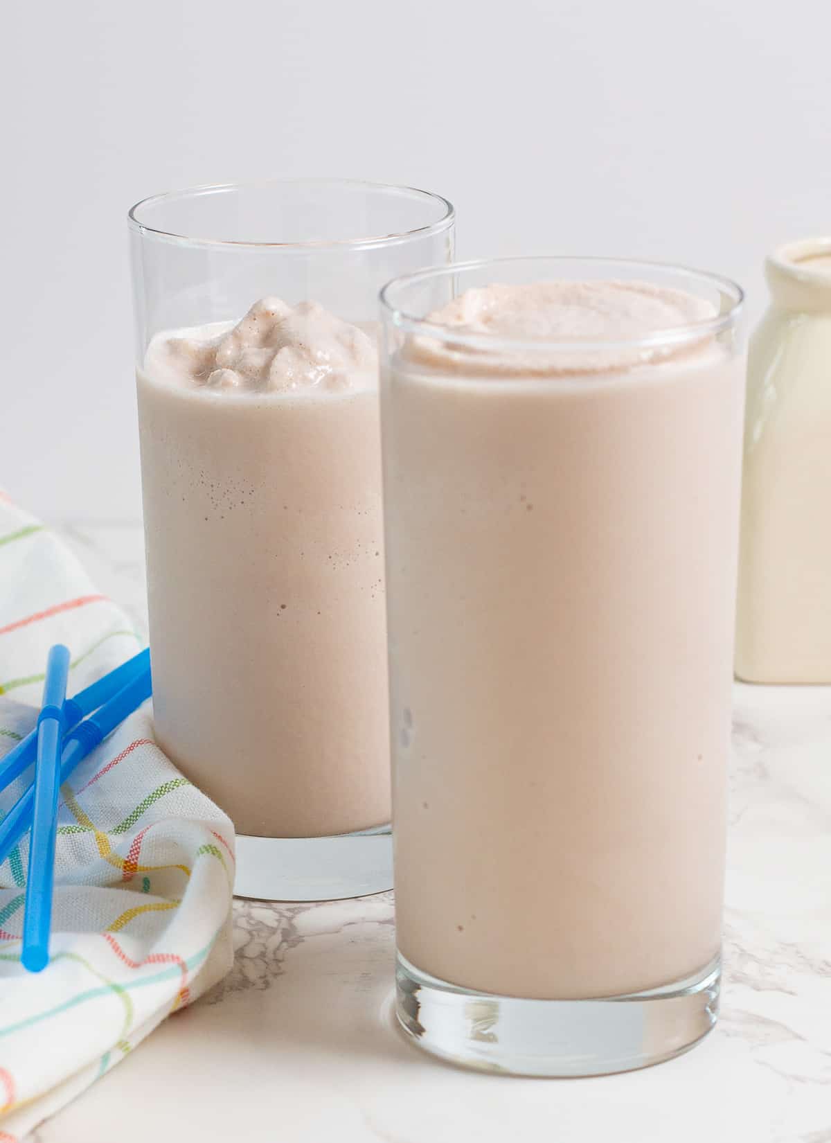 Two tall glasses filled with creamy chocolate milkshakes on a light surface, with a striped cloth and blue straws nearby.