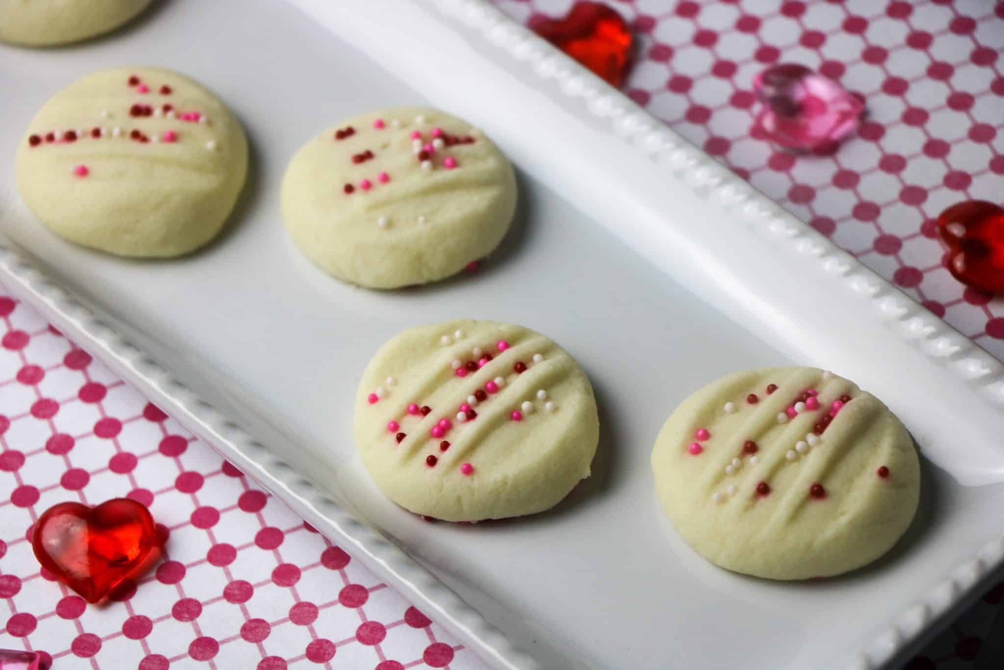 Round shortbread cookies with decorative fork marks and pink and white sprinkles, arranged on a white rectangular platter.