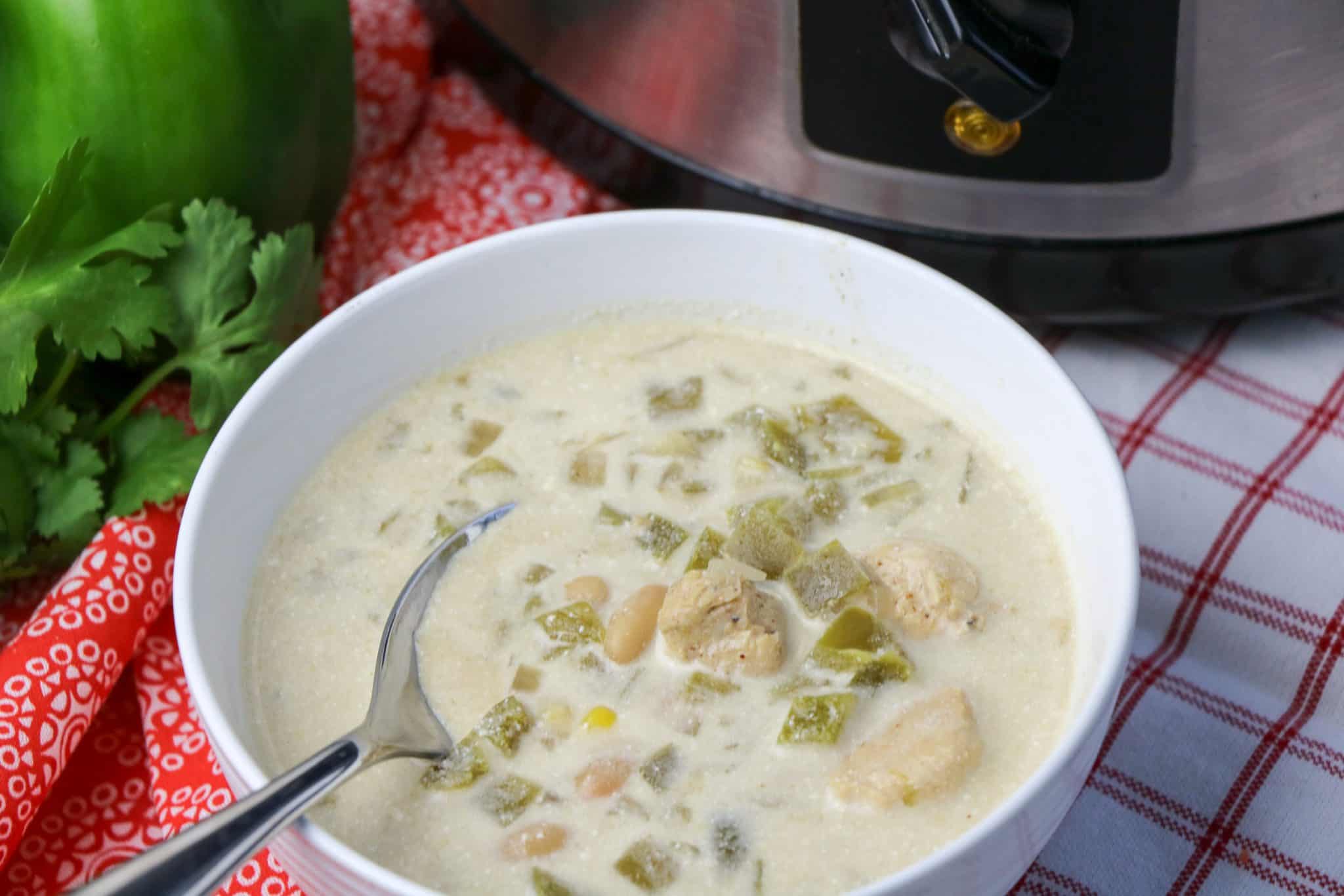 A bowl of creamy white chili with chicken, beans, and green chilies, with a spoon inside; slow cooker, cilantro, and green pepper are nearby.
