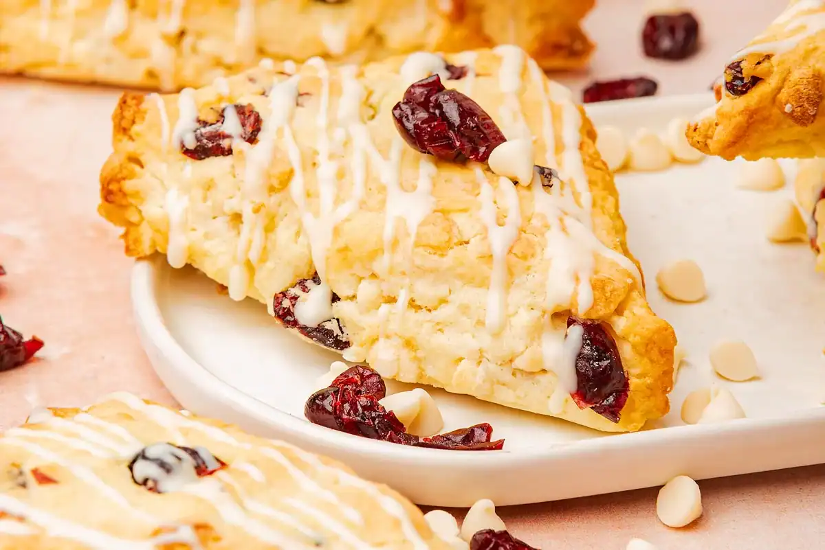 A triangular scone with white icing drizzle, visible dried cranberries, and white chocolate chips on a white plate.