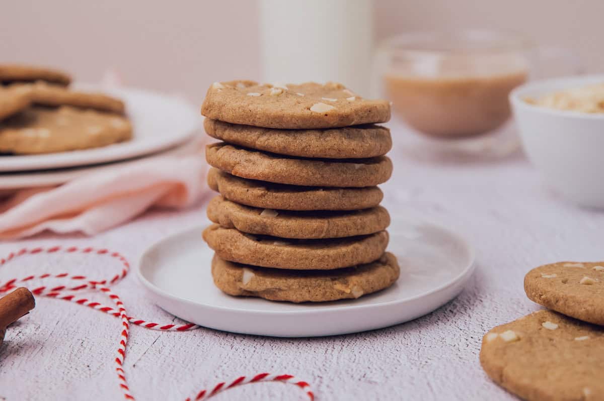 A stack of nine cookies with visible nut pieces sits on a white plate, surrounded by more cookies, a cup, and a glass of milk on a light-colored table.