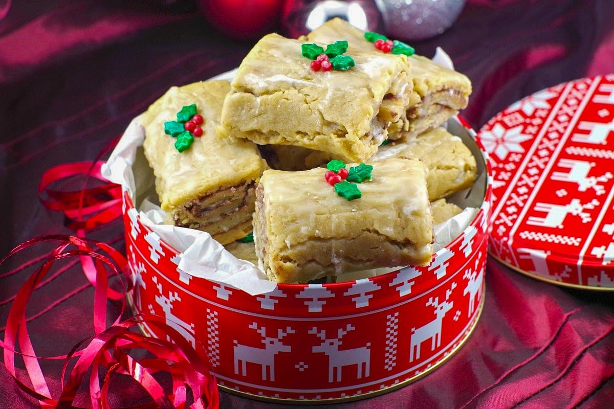 Rectangular iced pastries decorated with holly-shaped icing sit in a festive red tin with reindeer and trees, lined with white parchment paper.