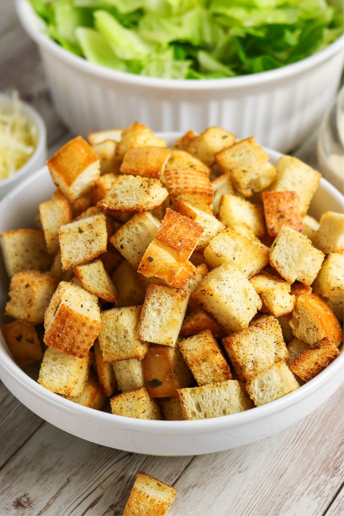 A white bowl filled with golden-brown croutons sits on a wooden surface, with a bowl of lettuce and a small dish of grated cheese in the background.