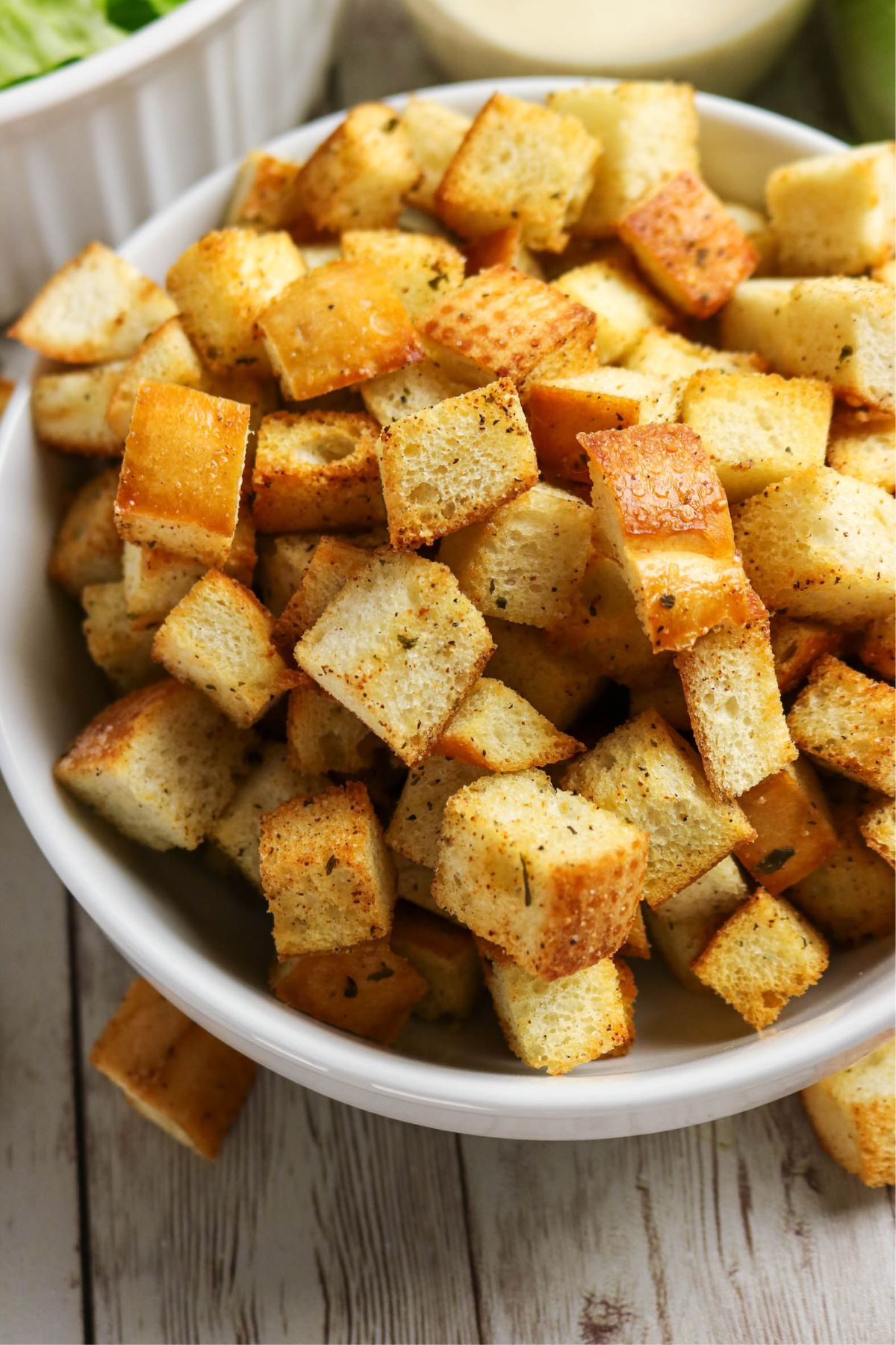 A white bowl filled with golden, seasoned croutons sits on a wooden surface.