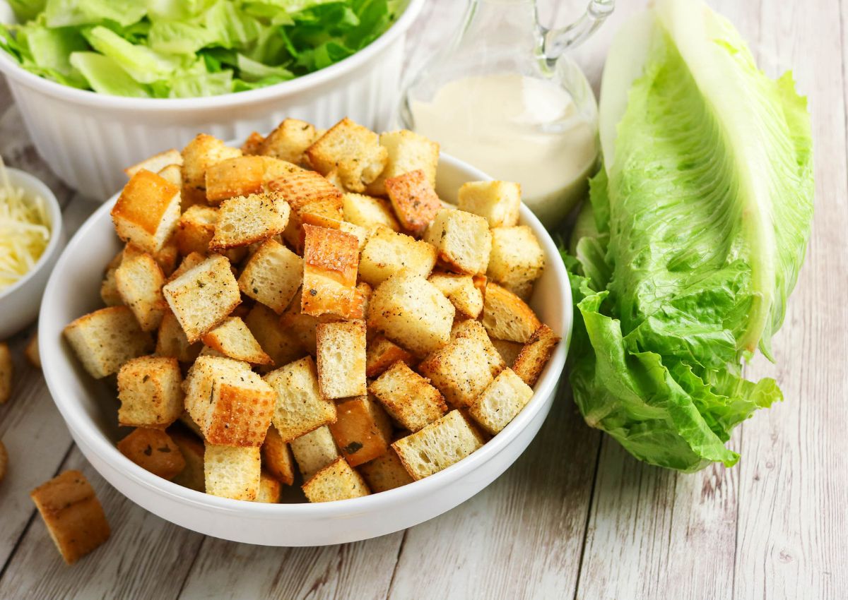A bowl of croutons sits on a wooden table next to fresh romaine lettuce, a bowl of greens, shredded cheese, and a small pitcher of salad dressing.