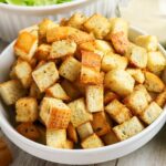A white bowl filled with seasoned, golden-brown croutons, with part of a salad and a dressing bowl visible in the background.