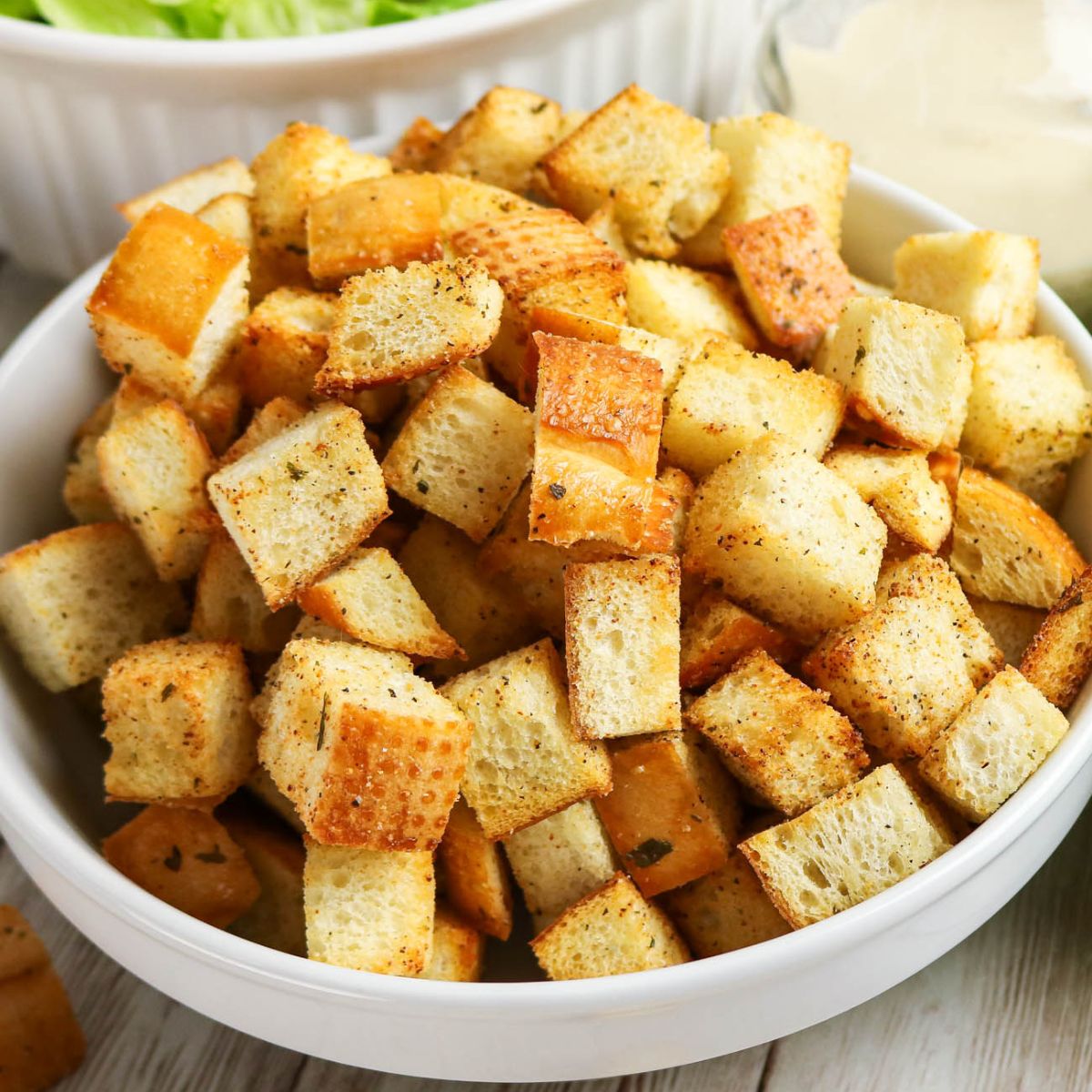 A white bowl filled with seasoned, golden-brown croutons, with part of a salad and a dressing bowl visible in the background.