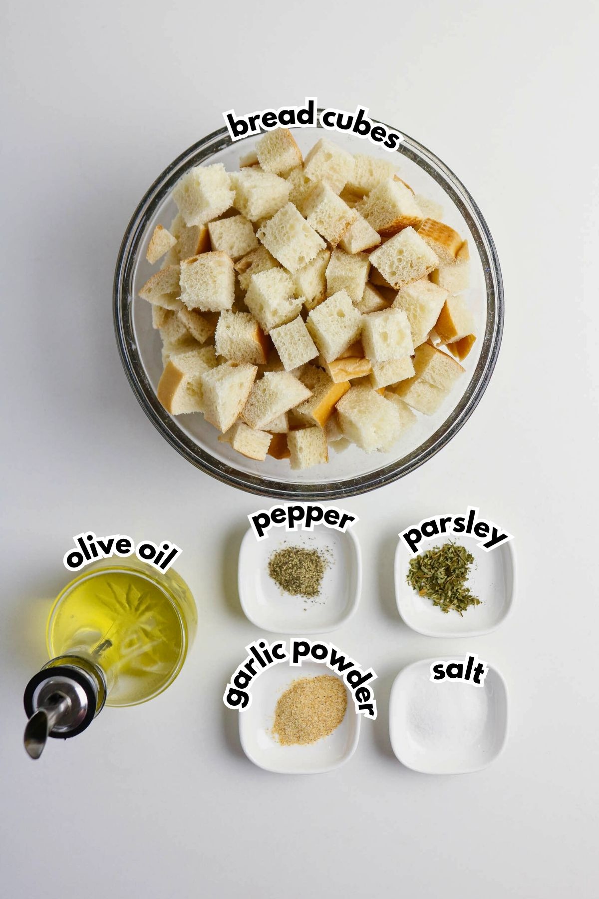 A bowl of bread cubes with small dishes of olive oil, pepper, parsley, garlic powder, and salt arranged on a white surface, each labeled with its name.