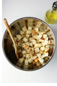 A metal bowl with cubed bread, black pepper, and a wooden spoon, next to a bottle of olive oil on a white surface.