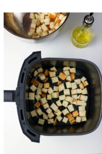 Cubed bread pieces are spread in an air fryer basket, with a metal bowl of more bread cubes and a bottle of oil nearby on a white surface.