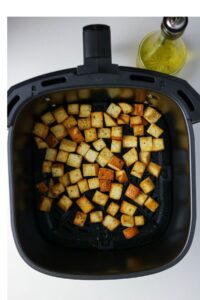 Cubed, seasoned croutons in an air fryer basket next to a bottle of olive oil on a white surface.