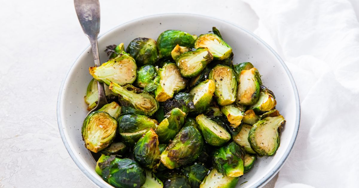A bowl of roasted Brussels sprouts with a fork, placed on a light-colored surface next to a white cloth.
