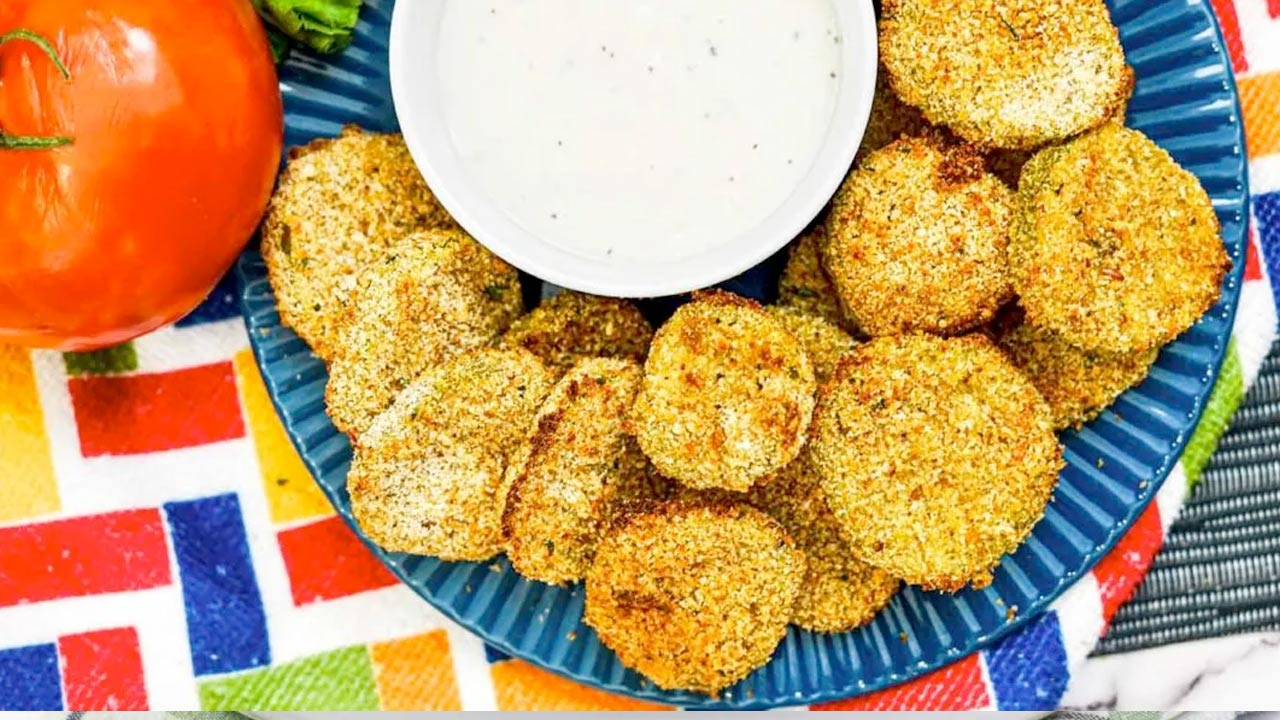 A blue plate with breaded fried green tomato slices arranged around a bowl of white dipping sauce, next to a whole tomato, on a colorful patterned cloth.