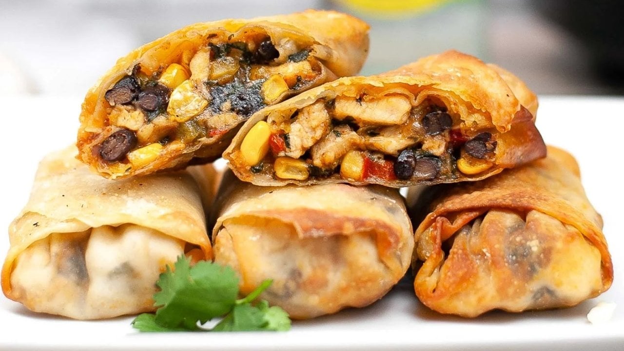 Four crispy, golden-brown egg rolls filled with black beans, corn, and vegetables are stacked on a white plate, with a sprig of cilantro nearby.