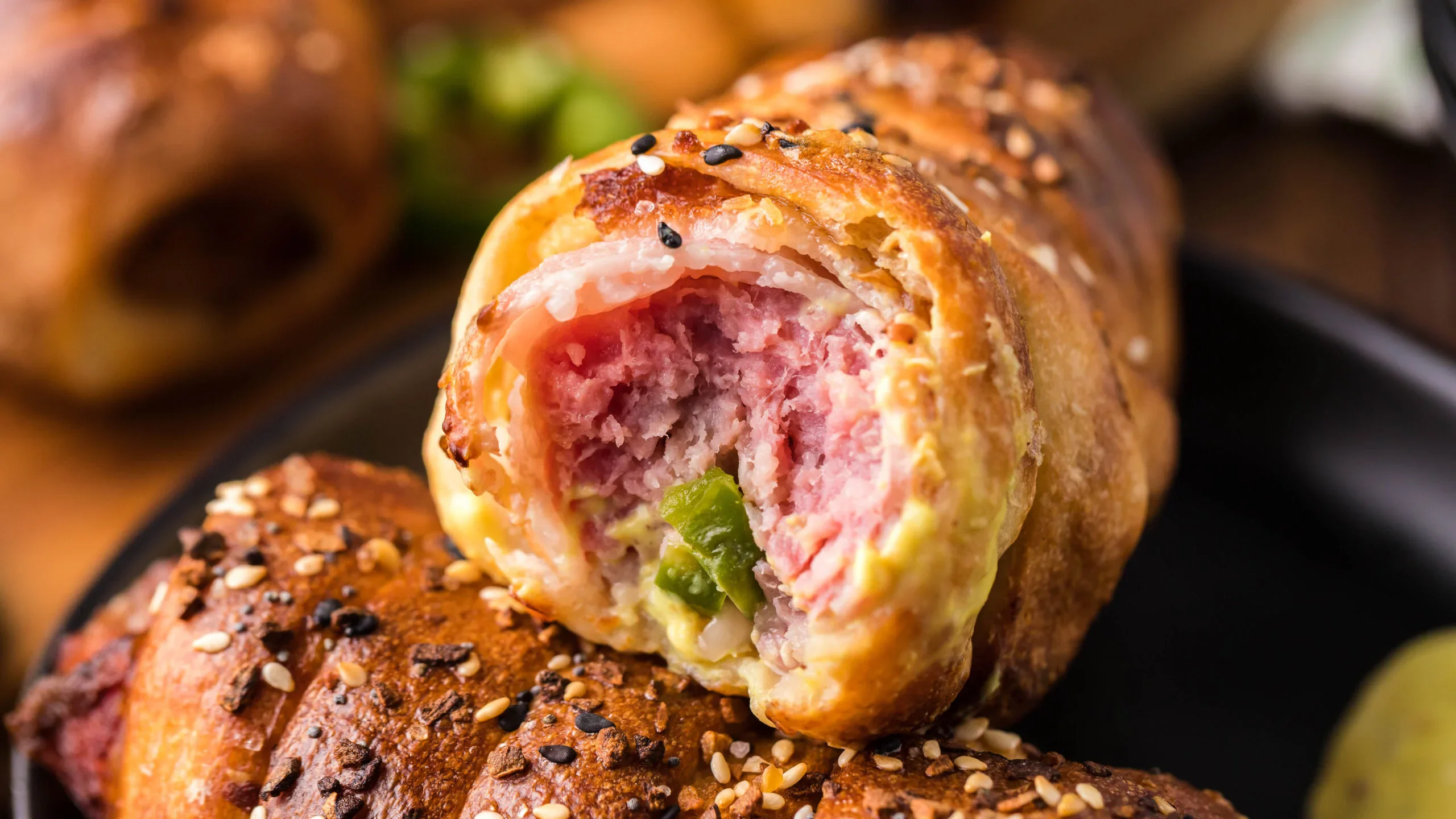 Close-up of a baked pastry filled with ham, cheese, and green peppers, topped with sesame and poppy seeds, with a bite revealing the filling.