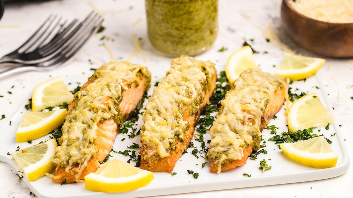 Three baked salmon fillets topped with melted cheese and herbs are arranged on a white plate, garnished with lemon wedges and chopped parsley.