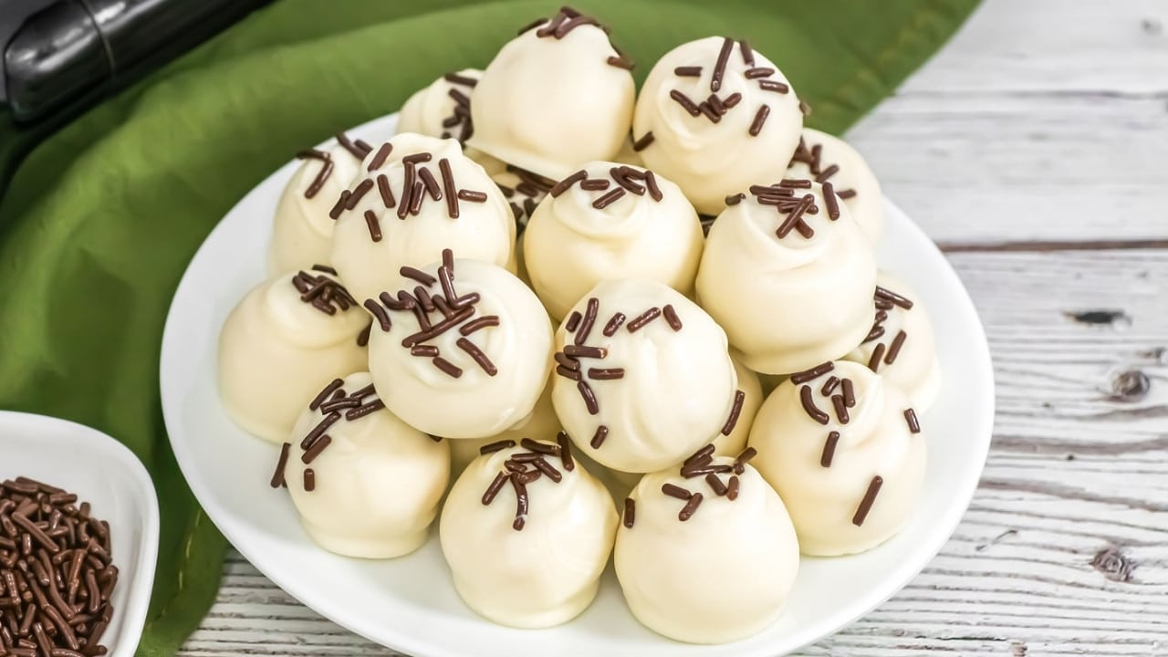 A white plate holds a stack of round white chocolate truffles topped with chocolate sprinkles, placed on a wooden surface next to a green cloth.