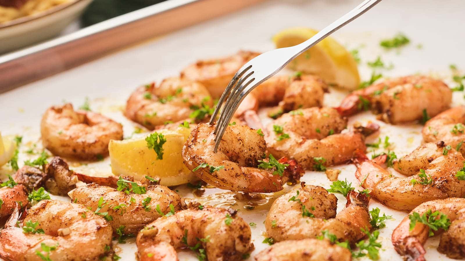 A fork picking up a seasoned, cooked shrimp from a tray of shrimp garnished with chopped herbs and lemon wedges.