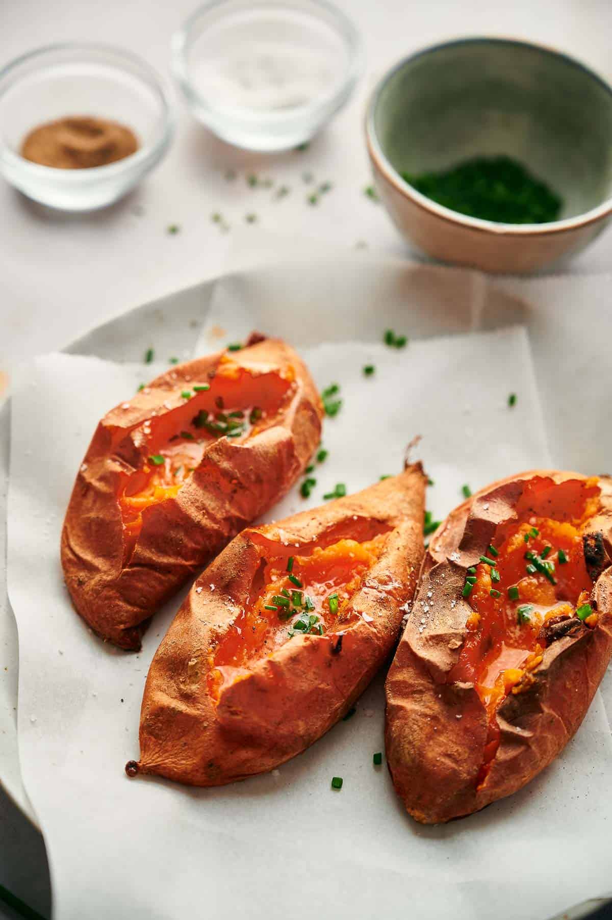 Three baked sweet potatoes topped with chopped chives are arranged on parchment paper, with small bowls of salt, pepper, and chives in the background.