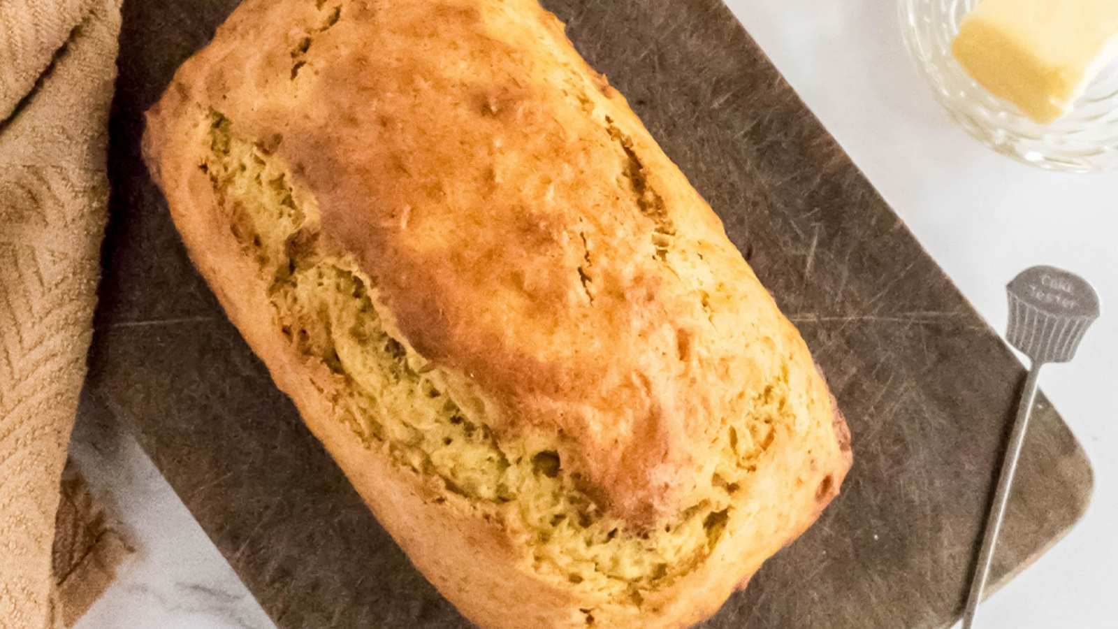 A loaf of golden-brown bread sits on a dark cutting board, with a butter dish and butter knife nearby.