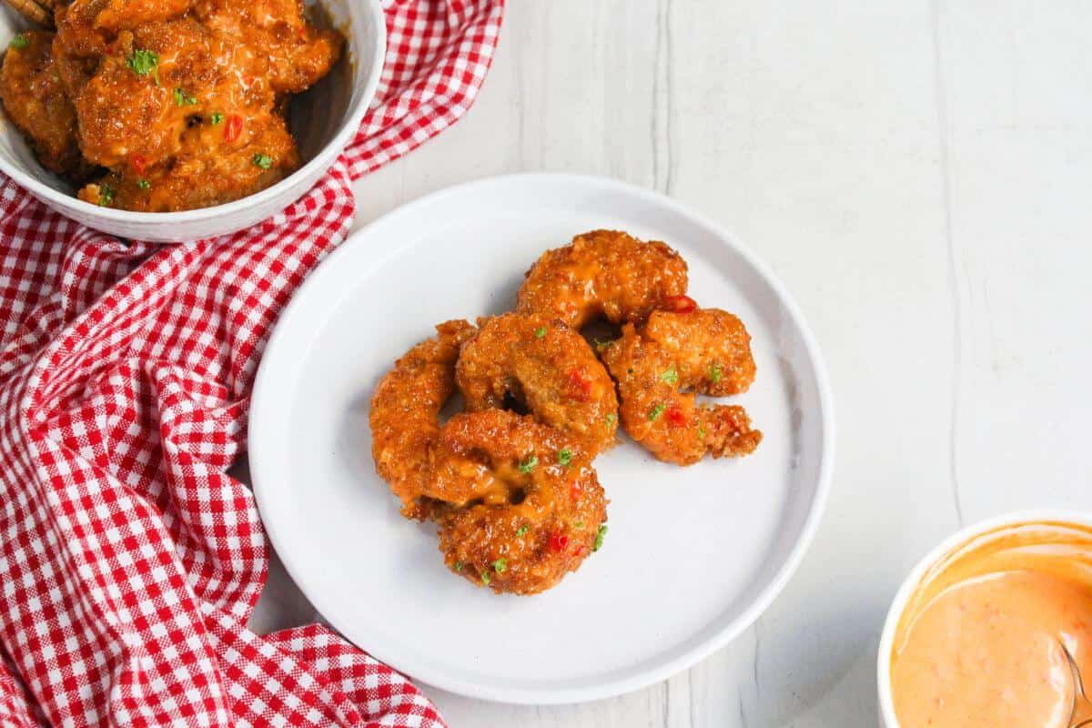 Five pieces of breaded fried shrimp on a white plate, with a bowl of sauce and a red checkered cloth nearby.