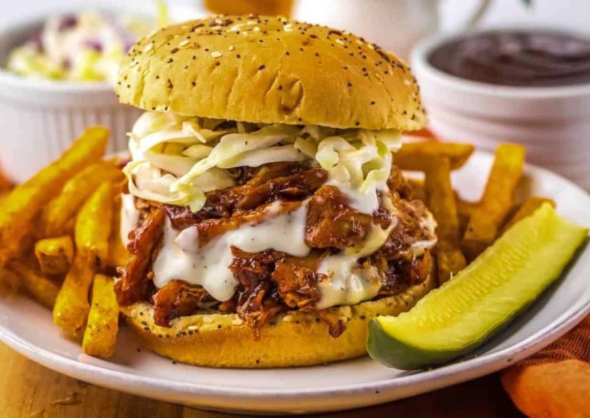 A plate with a barbecue pulled pork sandwich topped with coleslaw, served with French fries, a pickle spear, and side dishes in the background.