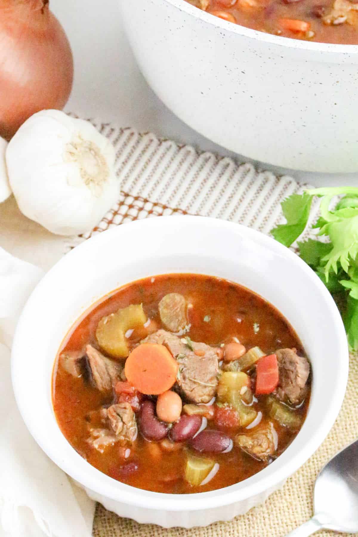 A bowl of hearty beef and vegetable soup with beans, carrots, celery, and tomatoes, placed on a table next to garlic, an onion, and a pot of soup.
