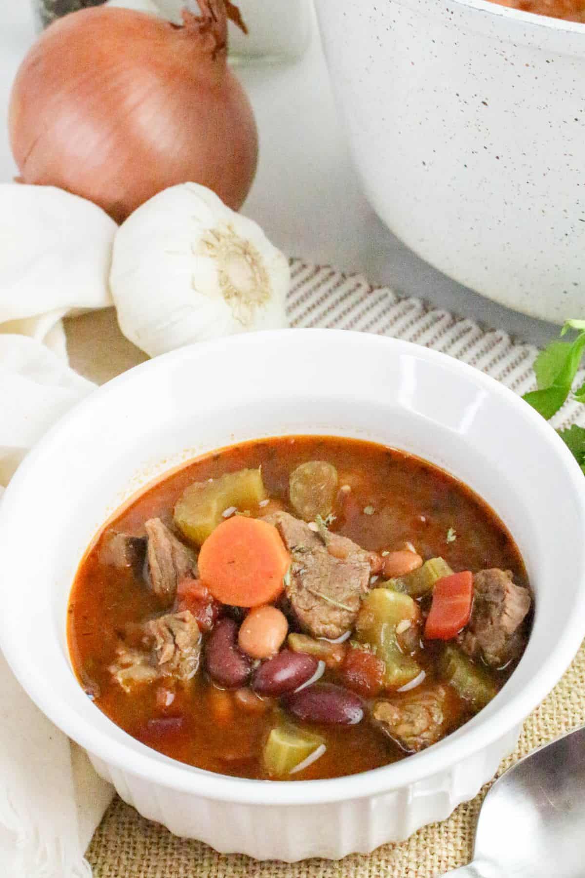 A white bowl of beef and vegetable stew with carrots, celery, beans, and chunks of meat. An onion, a garlic bulb, and a white pot are in the background.