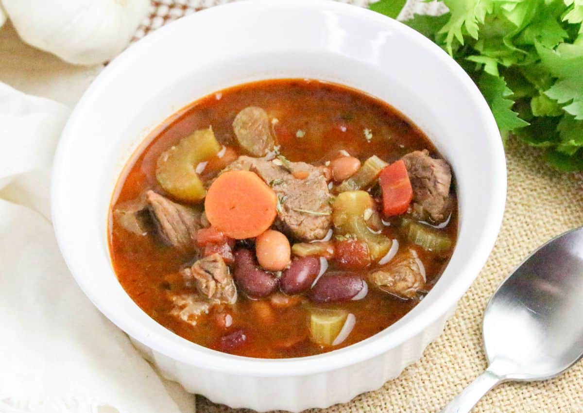 A white bowl filled with beef and bean soup, containing chunks of beef, kidney beans, pinto beans, celery, and carrot slices, placed next to a spoon and fresh cilantro.